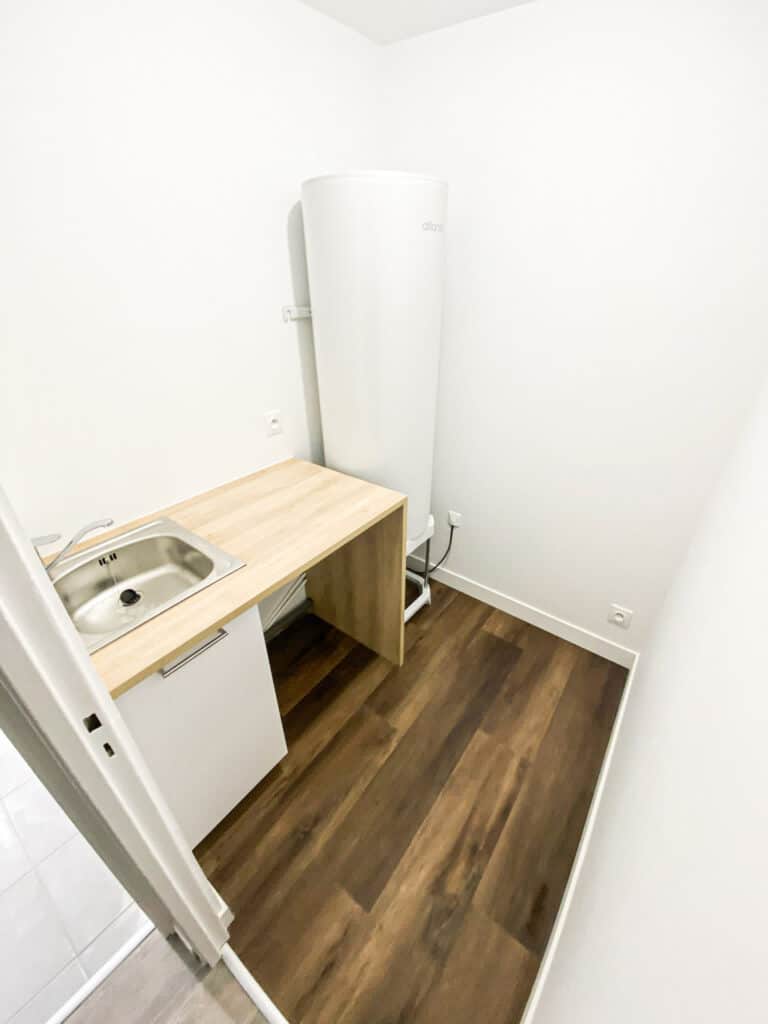 Buanderie compacte avec chauffe-eau Atlantic, évier inox sur plan de travail en bois clair, meuble bas. Sol stratifié foncé, murs blancs.