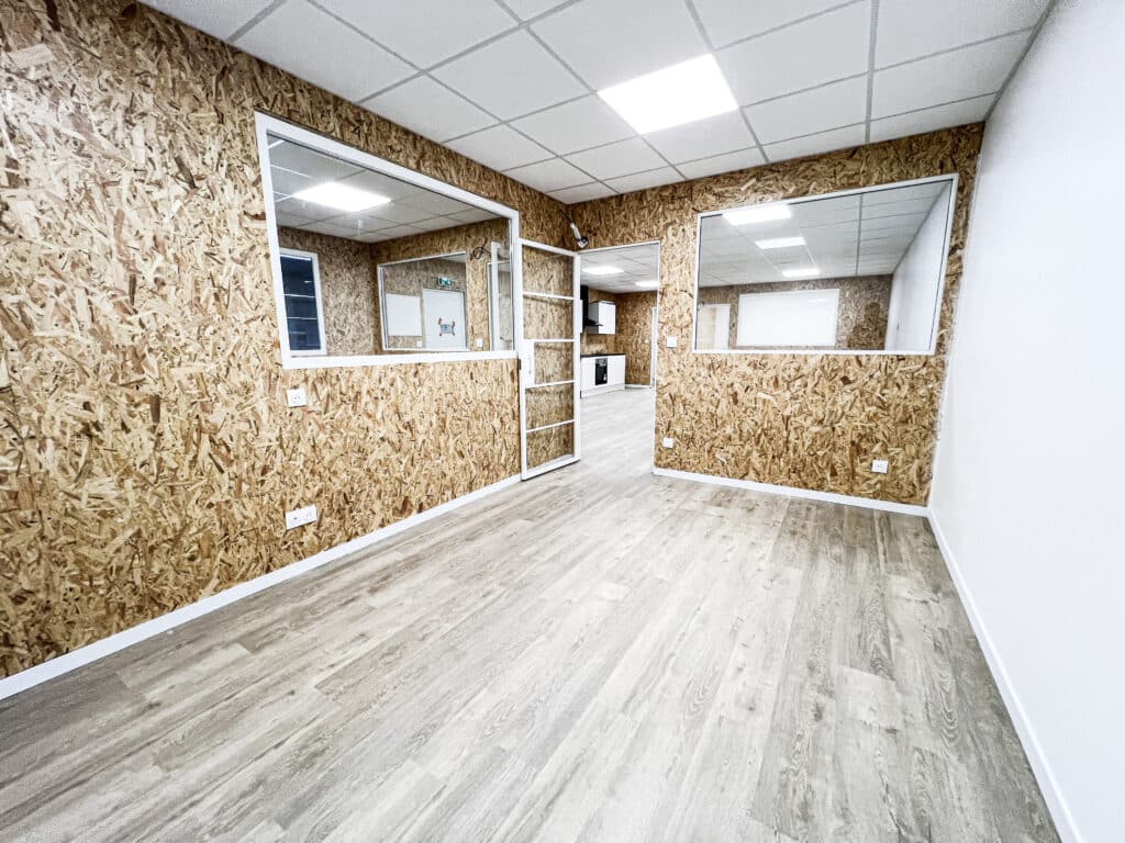 Bureau commercial moderne : murs OSB, plancher stratifié clair Espace commercial vide, murs en panneaux OSB, plancher stratifié clair, plafond quadrillé. Fenêtres intérieures et petite kitchenette équipée.
