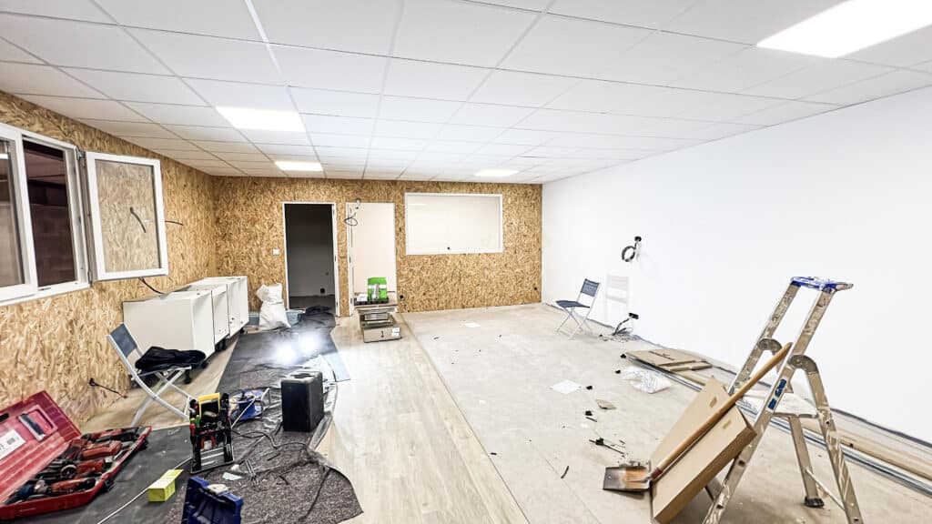 Pièce en rénovation avec murs en panneaux OSB et un mur blanc, plafond suspendu éclairé, sol stratifié. Outillage, échelle et fils électriques apparents suggèrent des travaux en cours.