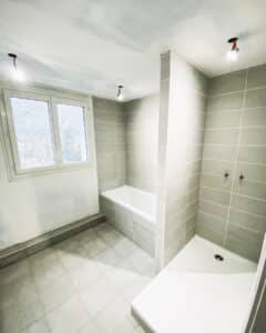 Salle de bain moderne en rénovation avec carrelage gris clair, baignoire blanche, douche à l'italienne et fenêtre lumineuse.