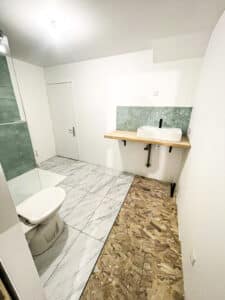 Salle de bain moderne en rénovation. Sol carrelé marbre avec douche, WC, et lavabo sur meuble bois avec dosseret hexagonal vert. Sol OSB à droite.