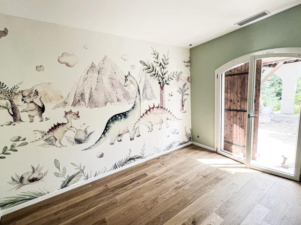 Chambre d'enfant dinosaures : fresque murale et parquet clair Chambre d'enfant lumineuse avec fresque murale de dinosaures, montagnes et arbres, parquet bois et mur vert sauge. Porte ouverte.