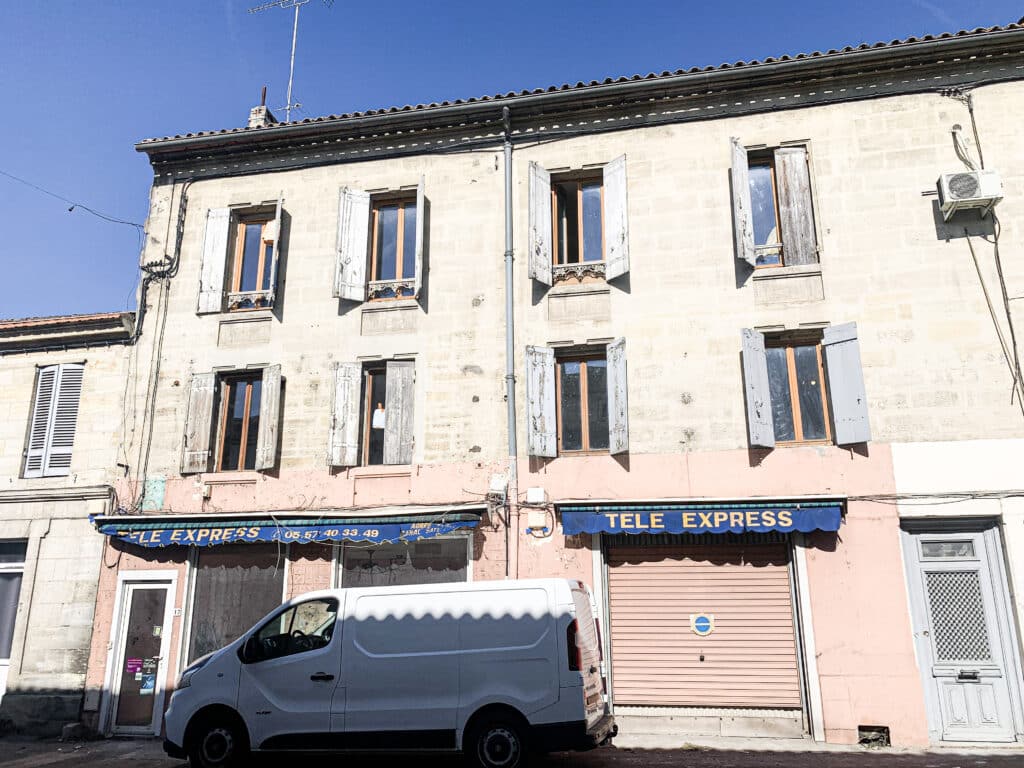 Façade de bâtiment européen avec volets vieillis, enseignes "TELE EXPRESS" et van blanc garé. Détails des câbles et AC unit.