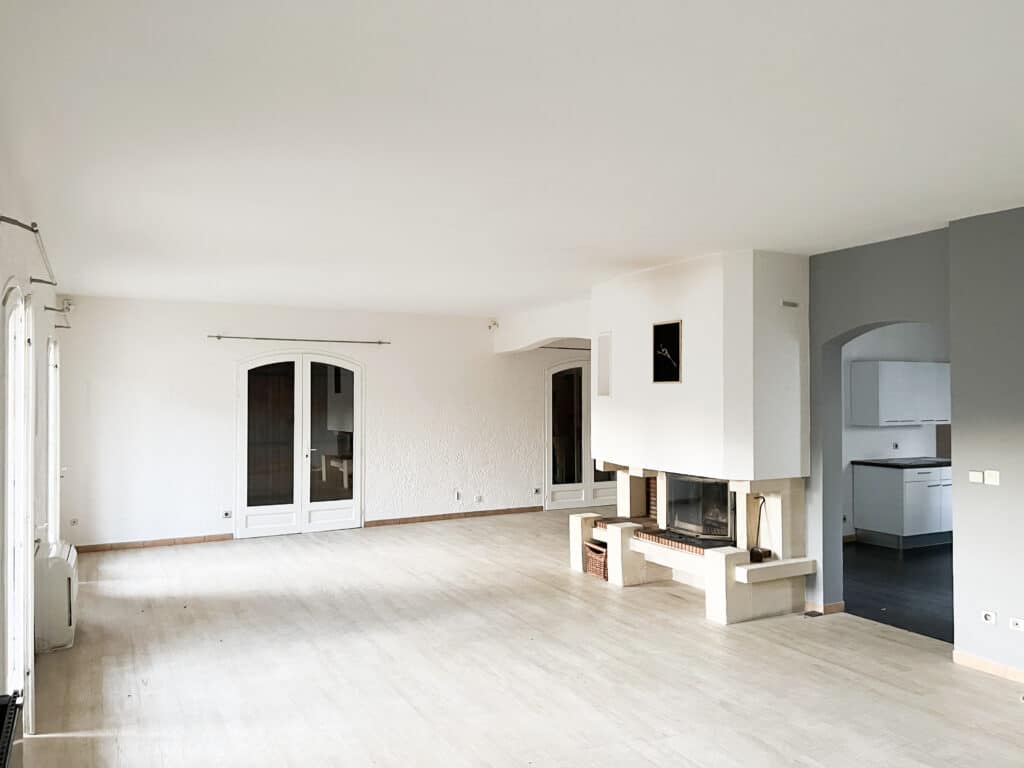 Grand salon vide, lumineux et rénové avec parquet clair. Cheminée moderne, portes-fenêtres, accès cuisine. Prêt à être aménagé.