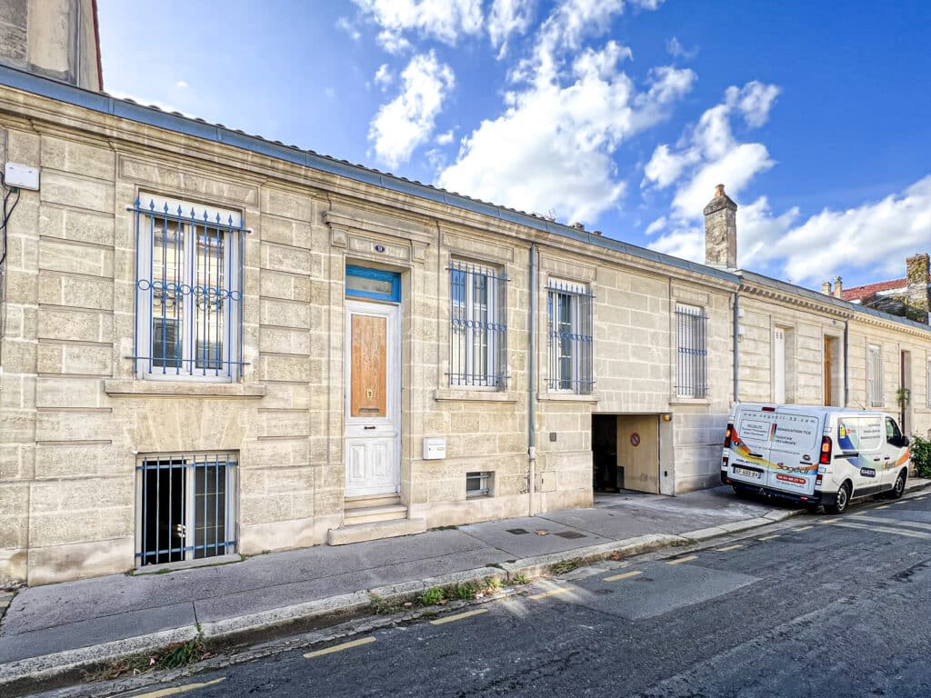 Façade en pierre d'une maison n°9 avec fenêtres à grilles bleues et porte blanche. Camionnette Sagecli-33.com de rénovation garée.