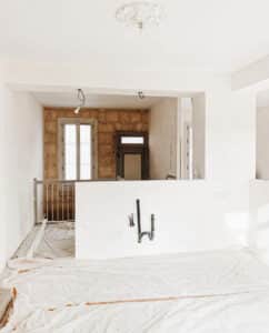 Rénovation d'appartement: murs blancs, plomberie cuisine/SDB, sol protégé. Mur de briques apparentes, fenêtre et porte ancienne en fond.