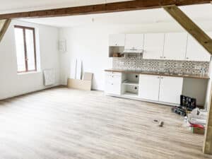 Cuisine ouverte moderne, meubles blancs, plan de travail bois. Crédence motifs beiges, parquet clair. Poutres apparentes. Pièce en rénovation.