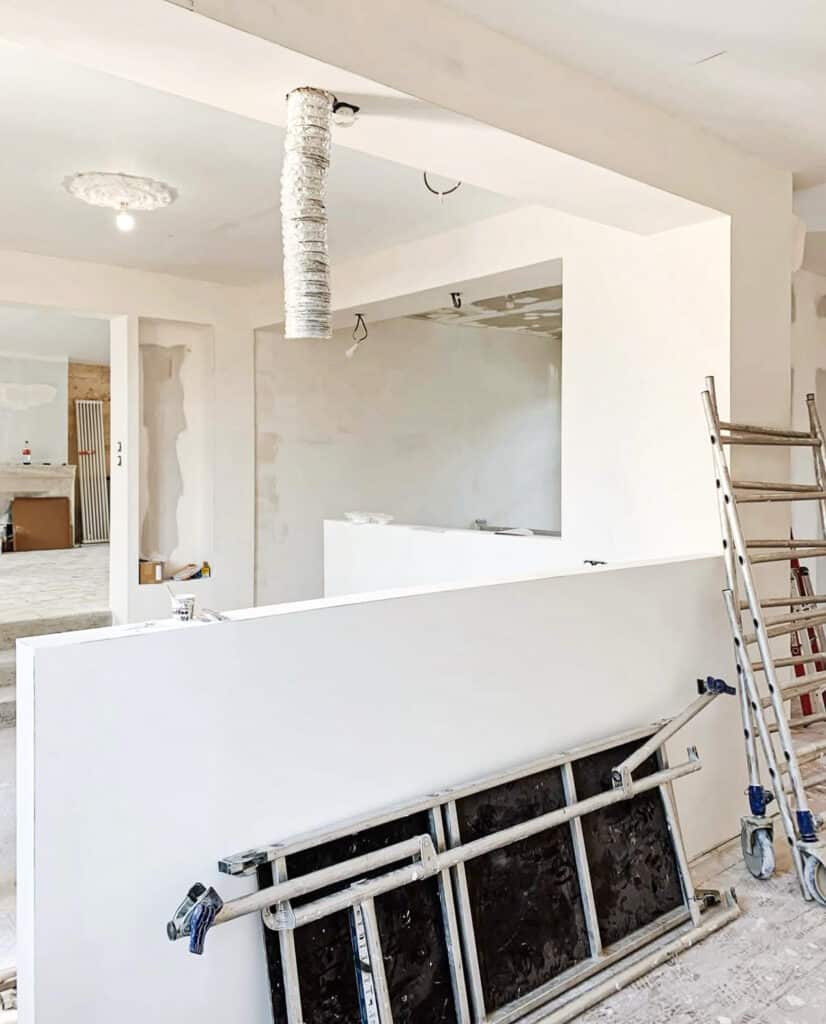 Intérieur d'une maison en rénovation avancée avec murs blancs, un lustre suspendu et un échafaudage métallique sur une cloison.