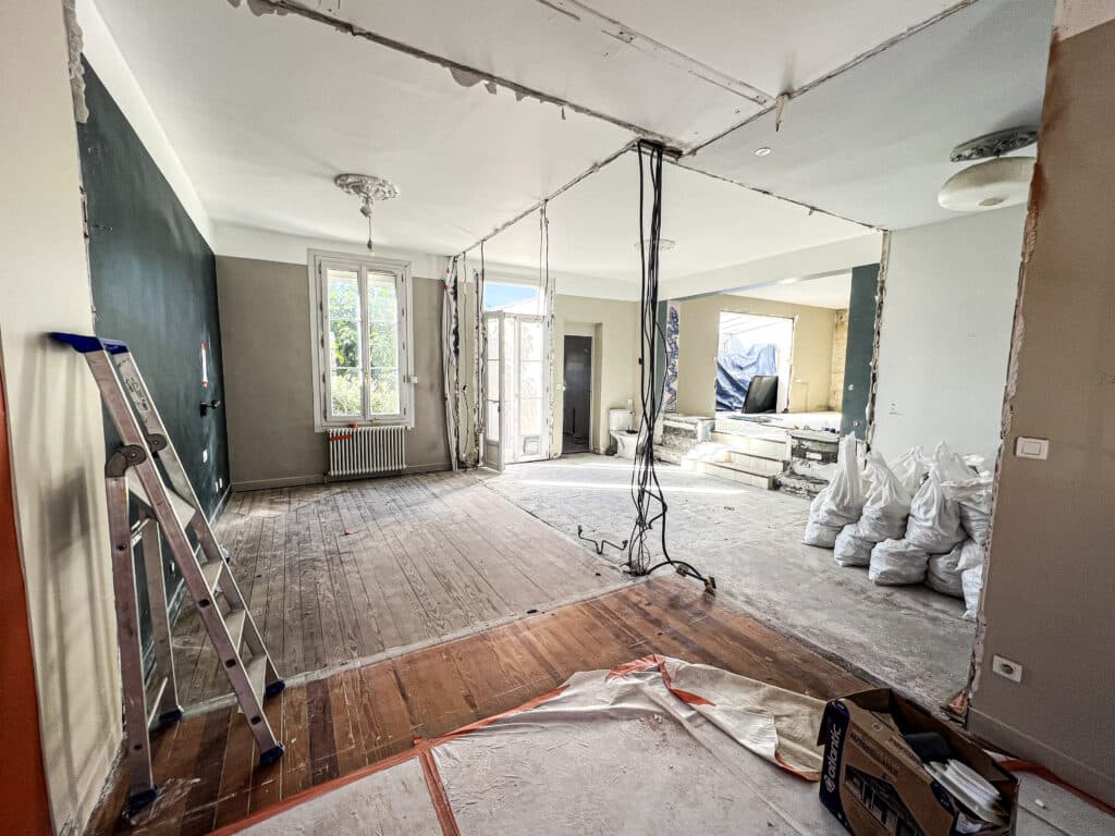 Chantier de rénovation d'un intérieur lumineux: fils électriques suspendus, murs démolis, parquet au sol, échelle et sacs de débris.