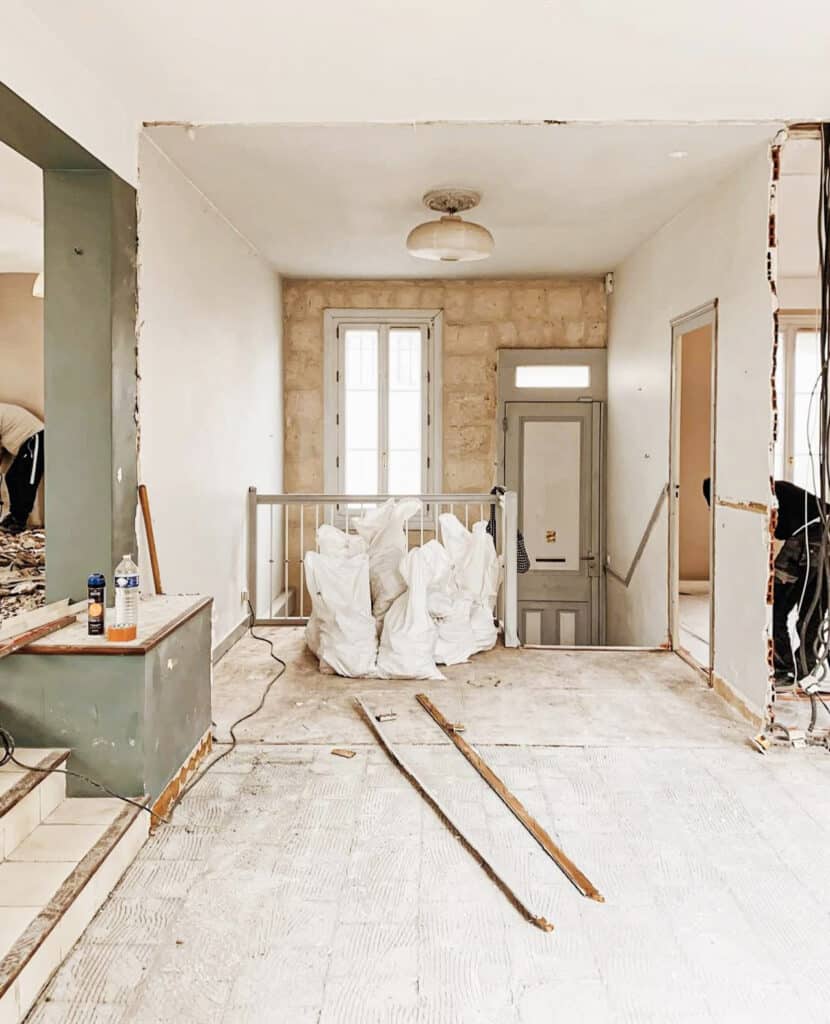 Rénovation maison : Démolition intérieure et préparation des travaux Intérieur d'une maison en rénovation, murs en partie démolis. Sol brut, sacs de gravats blancs et outils. Deux ouvriers travaillent près d'un escalier et de fenêtres.