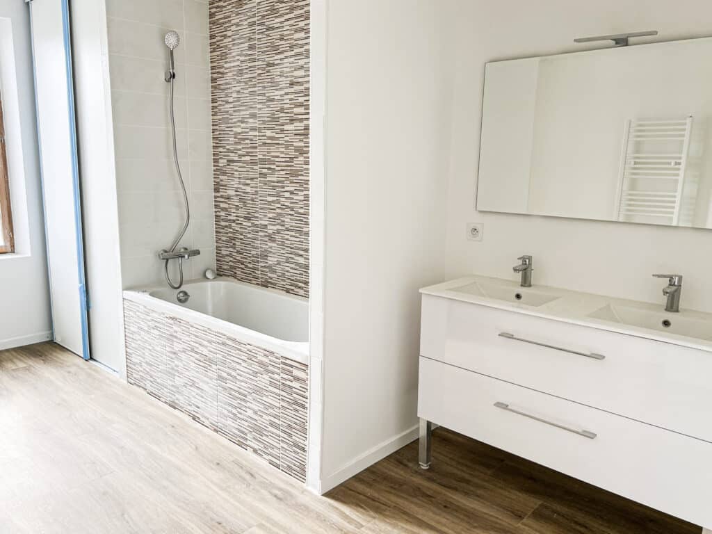 Salle de bain moderne : Baignoire, double vasque et miroir design Salle de bain moderne blanche avec baignoire carrelage mosaïque marron, meuble double vasque sous grand miroir éclairé. Sol aspect bois clair.
