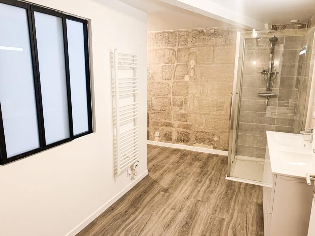 Salle de bain moderne: mur pierre apparente et douche design Salle de bain moderne: mur en pierre apparente, douche vitrée, lavabo blanc, radiateur sèche-serviettes, fenêtre noire, parquet bois.