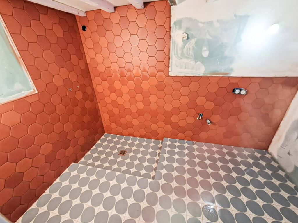 Salle de bain en rénovation, murs carrelés hexagonaux orange/terracotta, sol à motifs ovales gris. Plomberie et électricité apparentes.