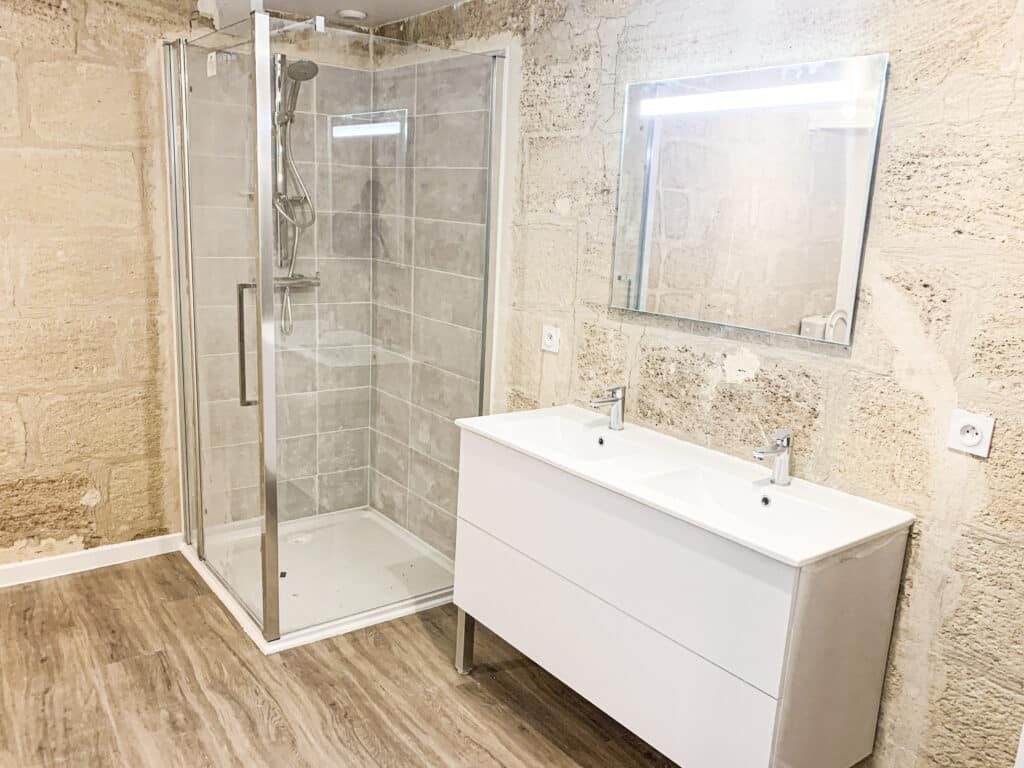 Salle de bain rénovée avec douche vitrée, double vasque blanche et miroir lumineux. Murs en pierres apparentes et sol imitation bois.