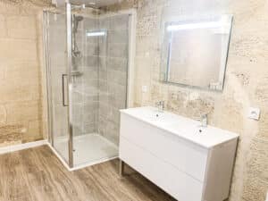 Salle de bain rénovée avec douche vitrée, double vasque blanche et miroir lumineux. Murs en pierres apparentes et sol imitation bois.