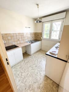 Cuisine ancienne avec évier double, plaque de cuisson noire, carrelage beige, sol vinyle marbré et fenêtre donnant sur immeuble extérieur. Dossiers et clés sur plan de travail.