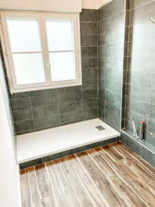 Salle de bain en rénovation : receveur de douche blanc, carrelage mural gris anthracite et sol imitation bois.