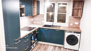 Cuisine moderne bleu sarcelle et bois clair, avec plaque induction, four encastré, évier noir, fenêtre et lave-linge. Texte visible : SHOT ON POCO X3 NFC.