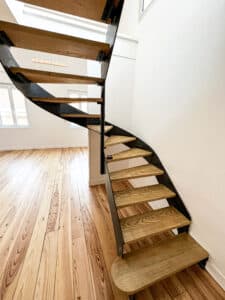 Escalier moderne à limon central noir avec marches en bois clair, ouvrant sur un intérieur lumineux avec plancher en bois.