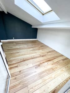 Grenier aménagé avec parquet clair, un mur bleu foncé contrastant, et une grande lucarne inondant la pièce de lumière naturelle.