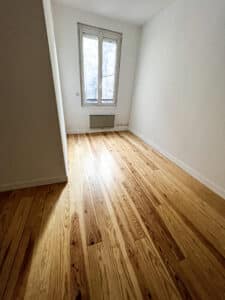 Pièce vide avec parquet bois clair neuf, murs blancs, un radiateur sous une fenêtre à petits carreaux et une ombre diagonale.
