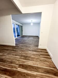 Intérieur d'une pièce vide avec sol imitation bois marron foncé et murs blancs, accès à un balcon par porte-fenêtre.