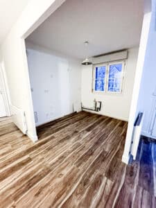 Pièce vide avec sol stratifié imitation bois foncé, murs blancs, fenêtre donnant sur un bâtiment bleu, et radiateurs en attente.