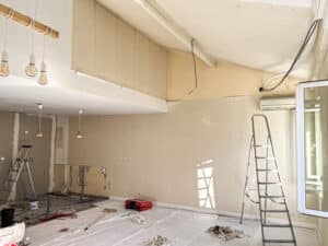 Chantier de rénovation intérieure avec murs en cours de préparation, échelle, luminaires suspendus en bois et revêtements de sol protégés.