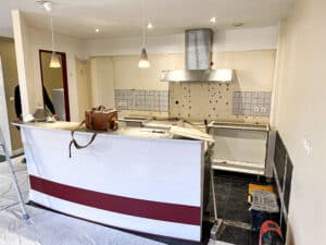 Rénovation cuisine : comptoir blanc et bordeaux, murs démontés, hotte inox, ouvrier en arrière-plan travaillant.