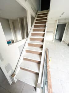 Escalier en cours de rénovation, contremarches provisoires en aggloméré, murs en placo brut, carrelage au sol. Matériaux de chantier visibles.