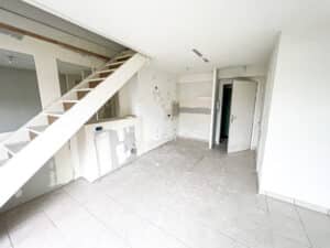 Intérieur d'une pièce en rénovation, murs blancs à finir, escalier blanc, et sol carrelé gris sale.