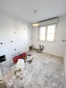 Chambre en rénovation avec sol décapé, prises apparentes, arrivée d'eau/tuyauterie encastrée, outils et sac de matériaux.
