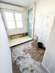 Salle de bain en rénovation : sol boueux et dégradé, douche neuve partiellement installée près d'une fenêtre, murs ouverts et carrelage partiel.