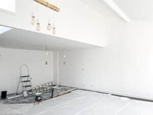 Intérieur de pièce neuve ou rénovée, murs blancs, sol protégé par bâches, échelles et seaux de peinture indiquent des travaux en cours.
