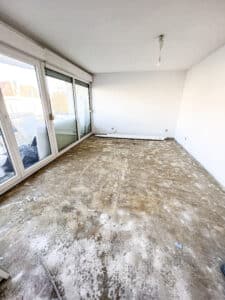 Pièce en rénovation avec sol brut après retrait de revêtement, murs blancs, et grandes baies vitrées donnant sur un balcon.
