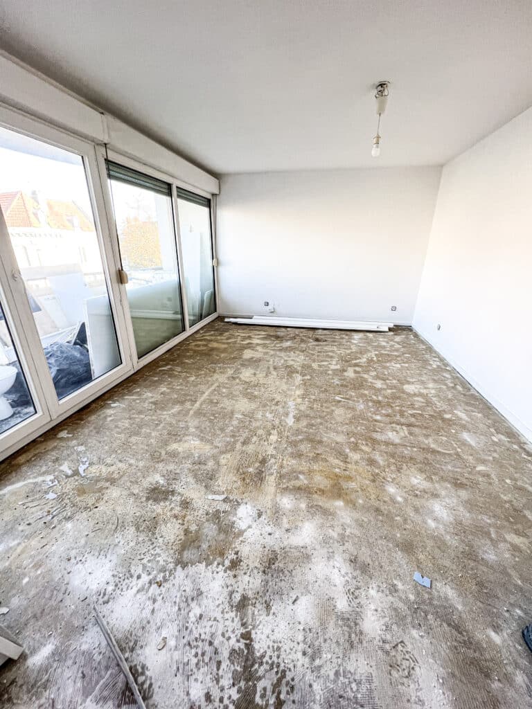 Pièce en rénovation avec sol brut après retrait de revêtement, murs blancs, et grandes baies vitrées donnant sur un balcon.