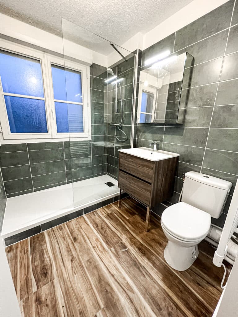 Salle de bain moderne avec douche vitrée, meuble vasque bois foncé, toilettes blanches et carrelage mural gris-vert foncé.
