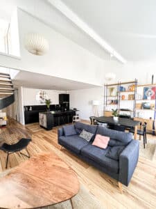 Salon moderne lumineux avec parquet bois clair, canapé gris foncé, table basse ronde bois, cuisine noire et étagère murale. Affiche "LIBERTÉ ÉGALITÉ FRATERNITÉ".