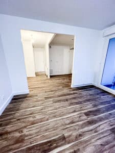 Pièce vide aux murs blancs et plafond texturé, sol imitation bois brun foncé, menant à un couloir et une fenêtre.