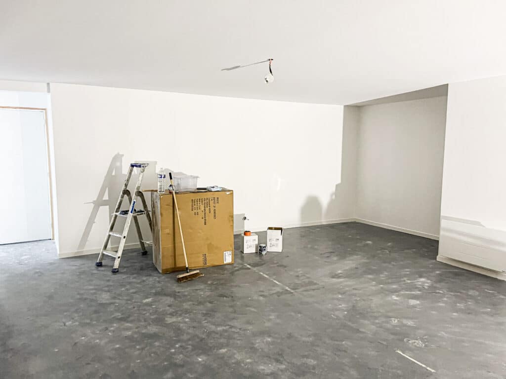 Pièce vide avec murs blancs fraîchement peints, sol gris, échelle, balai et carton de livraison marqués "BUFFET 2P PERROUET".