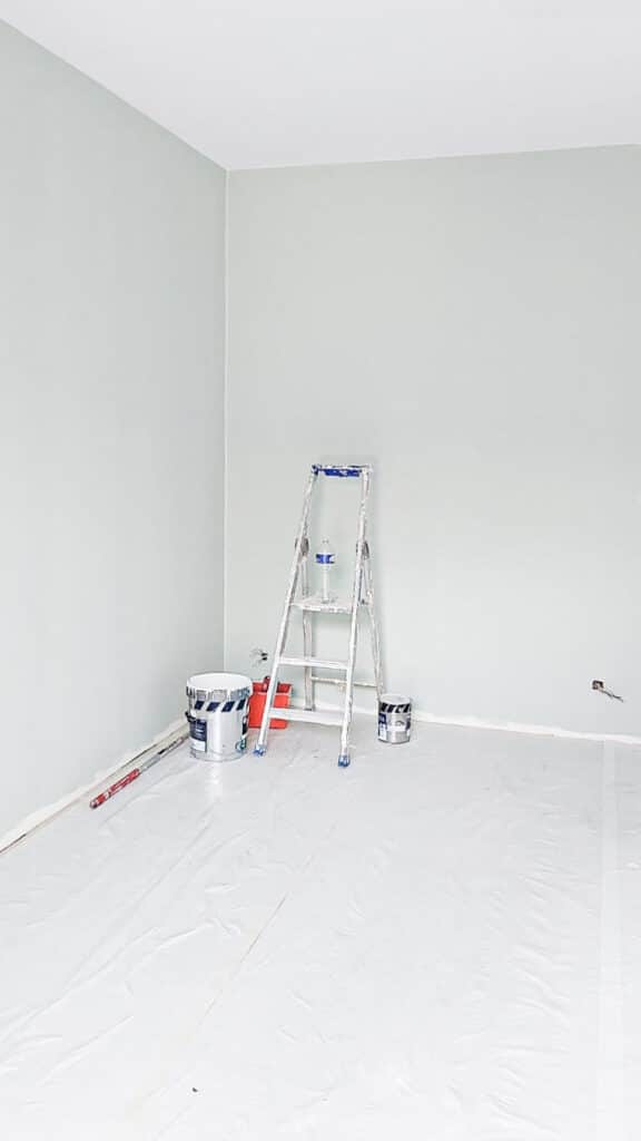 Chambre en cours de peinture, murs vert pâle, sol bâché de blanc, escabeau métallique avec bouteille d'eau, pots de peinture visibles.