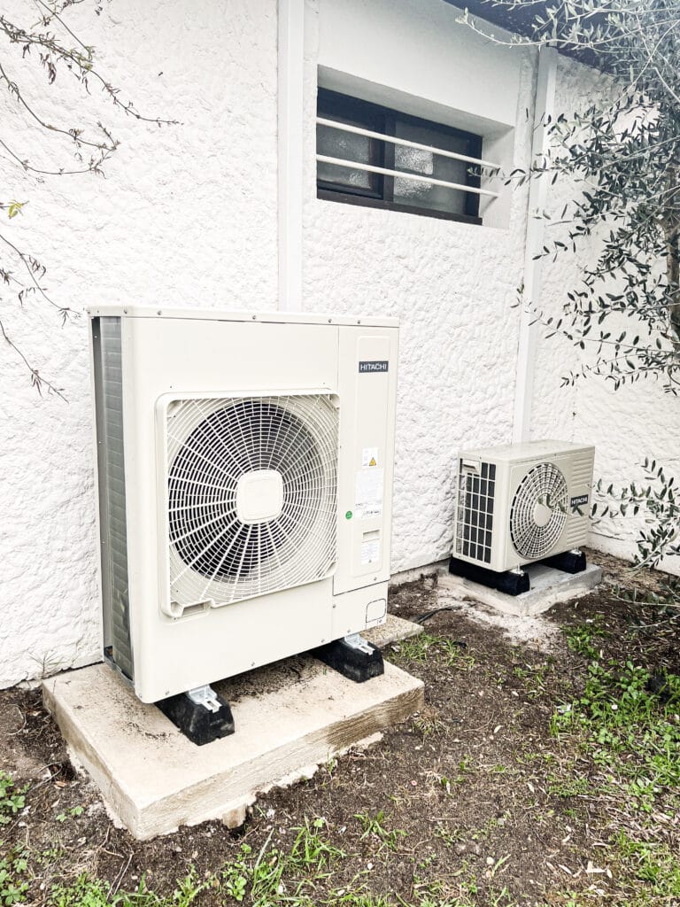Installation extérieure Hitachi : Pompe à chaleur et climatiseur. Deux unités extérieures HITACHI (pompe à chaleur/climatiseur) sur dalles de béton contre un mur blanc texturé, près d'un olivier.