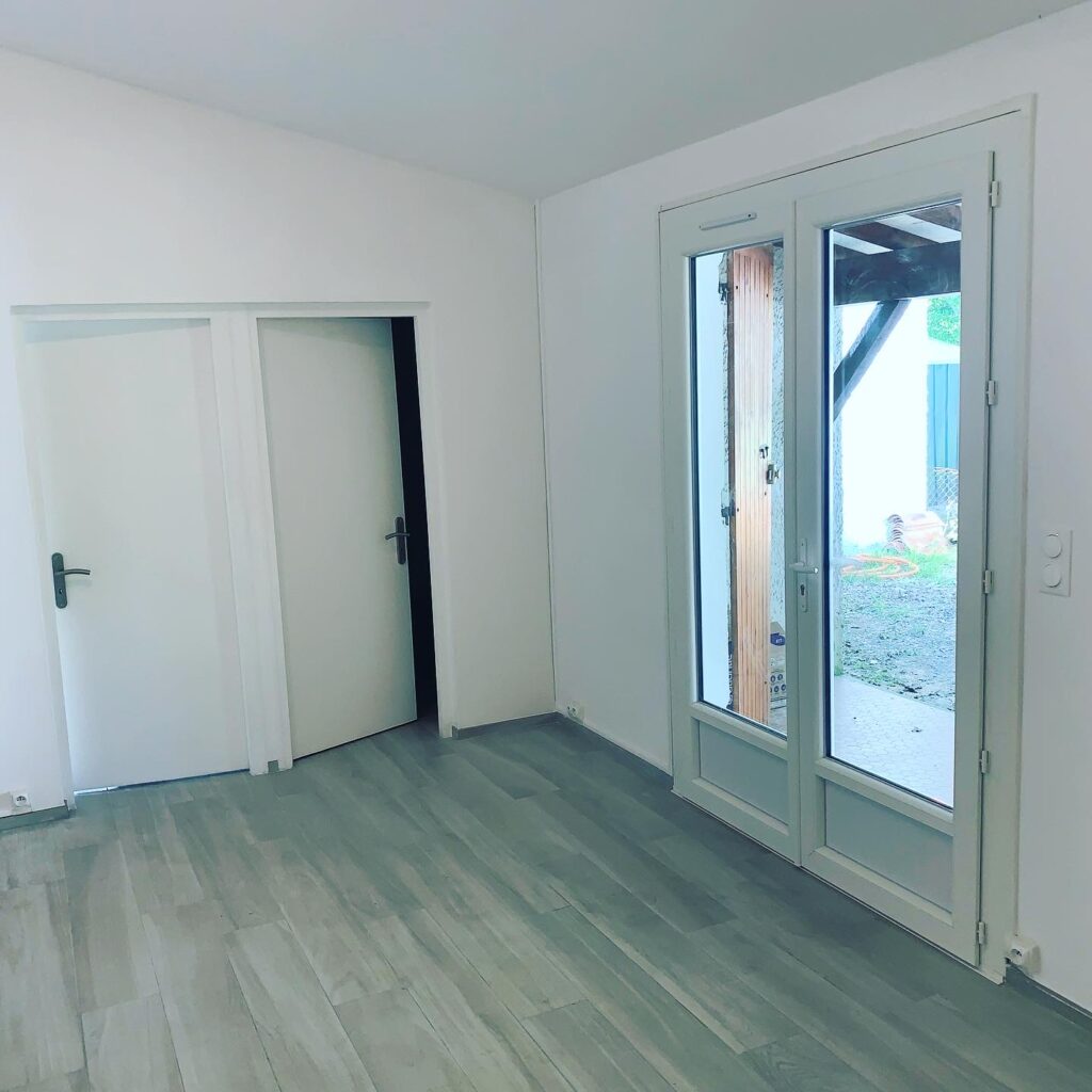 Pièce Lumineuse avec Parquet Gris et Grande Baie Vitrée Pièce vide lumineuse avec parquet gris, deux portes blanches et une grande baie vitrée ouvrant sur un jardin.